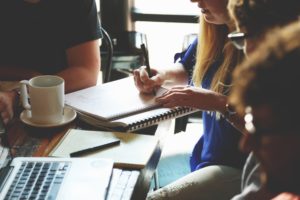 What is QA Testing (picture shows a meeting table with two people sitting at it. One is wearing a black t-shirt with an arm visible. The other is a blonde white woman wearing a blue shirt and jeans, writing on a notebook)