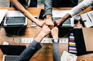 Agile QA Team (picture shows five hands doing fist bumps; one is Latina, three are white, and one is Black. They are sitting at a table with laptops and power cords on it)