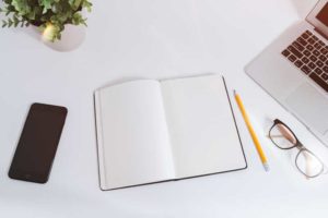 Writing Test Cases (picture depicts a white desk with a blank iPhone, notebook, pencil, pair of glasses, and Macbook laptop)