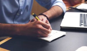 Prioritizing Bug Fixes (picture shows the bottom half of a person wearing a blue work shirt, sitting at a desk writing in a notebook)