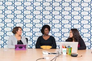 Women in Tech (picture shows a blue wallpaper background, with three women of color sitting at a table with laptops)